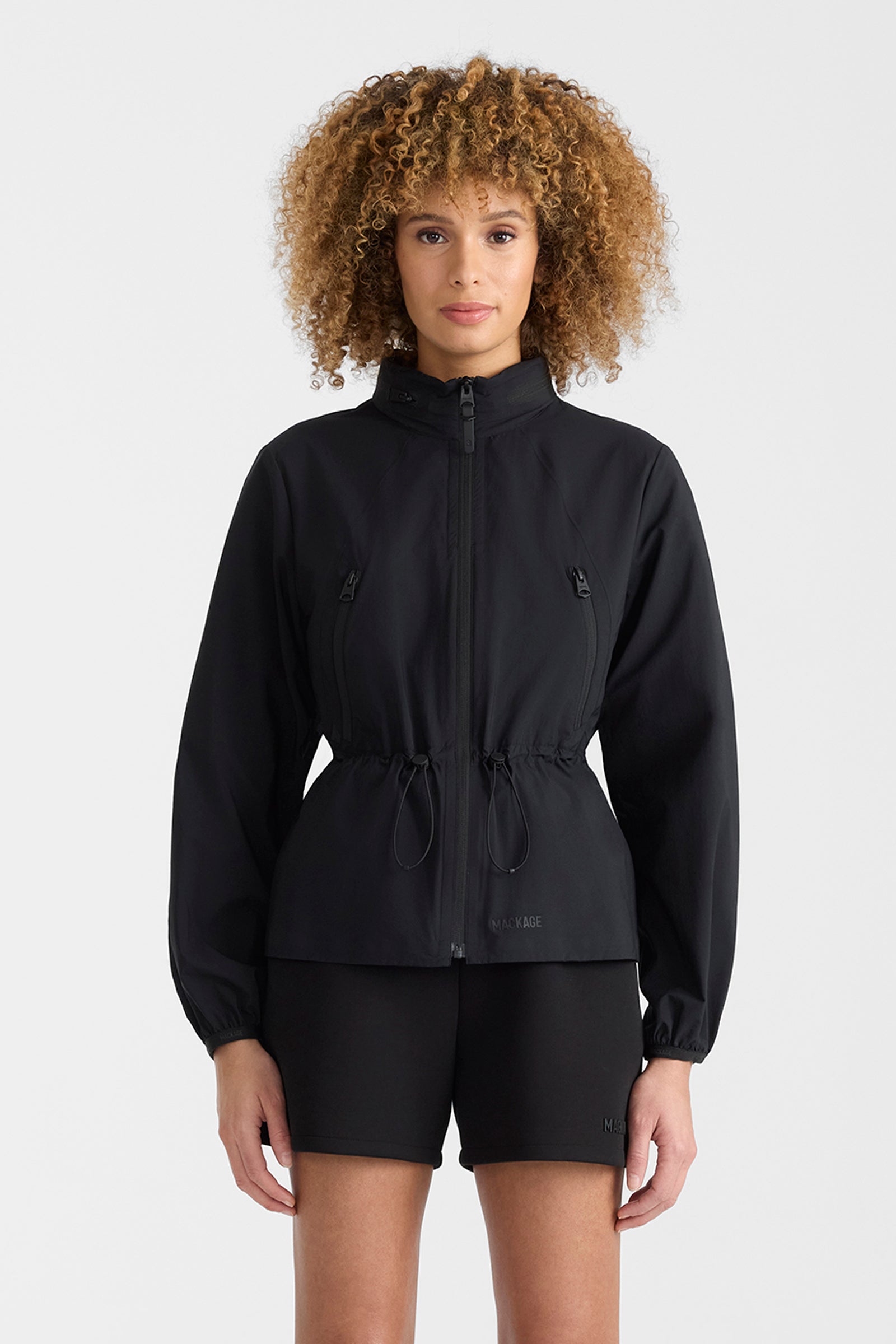 Woman wearing a black lightweight rain jacket with black shorts on a white background.