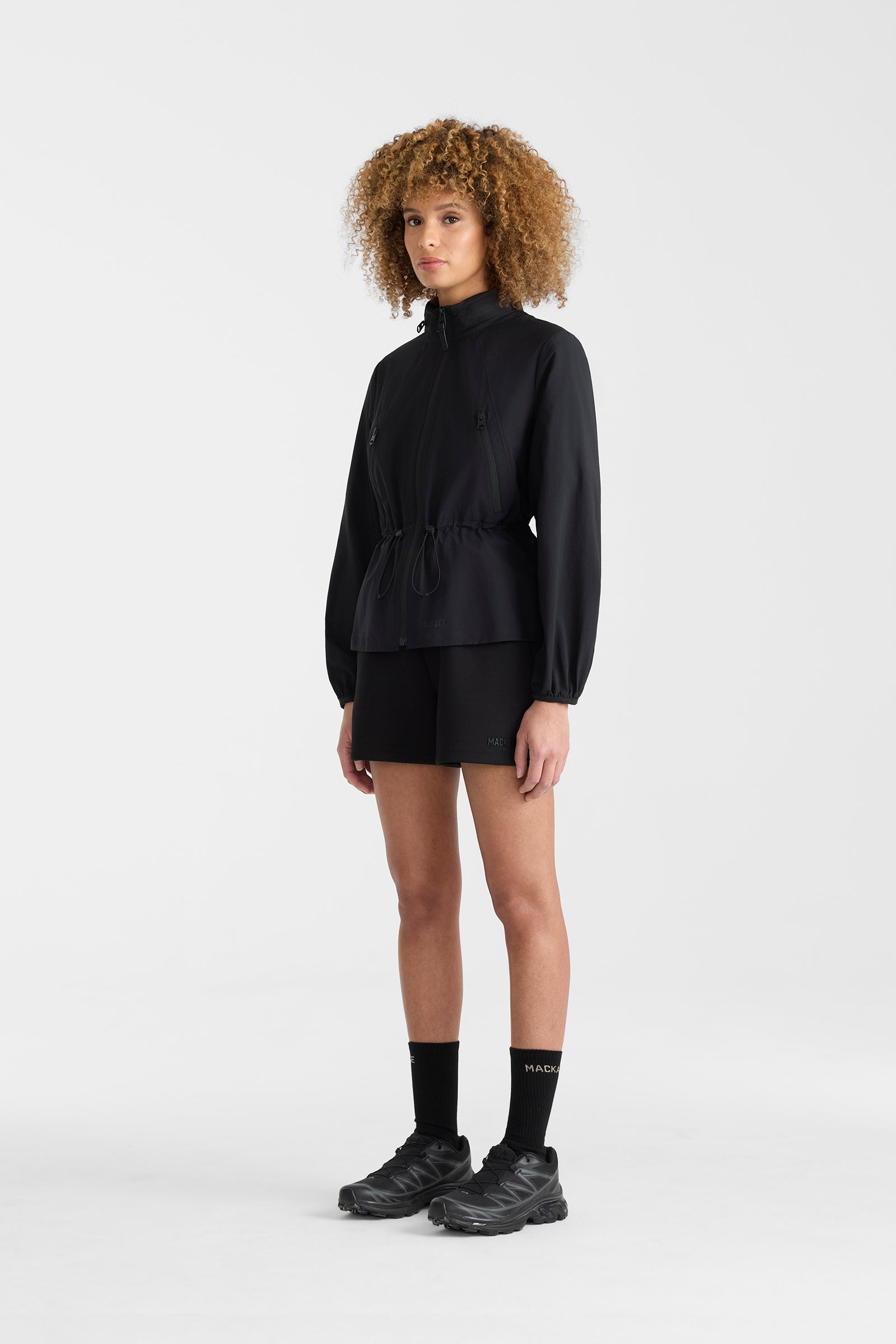 Woman wearing a black lightweight rain jacket with black shorts on a white background.