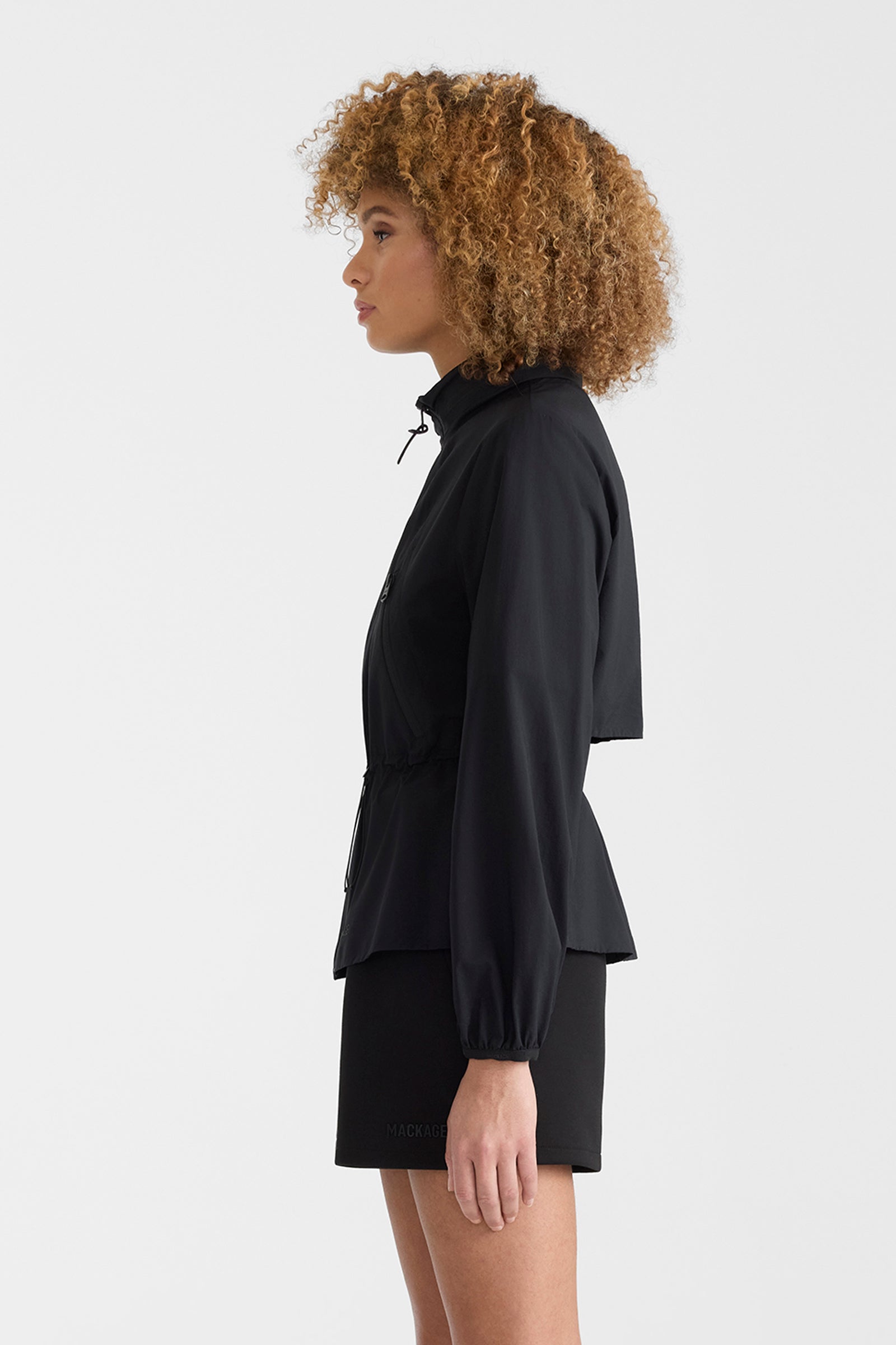 Woman wearing a black lightweight rain jacket with black shorts on a white background.