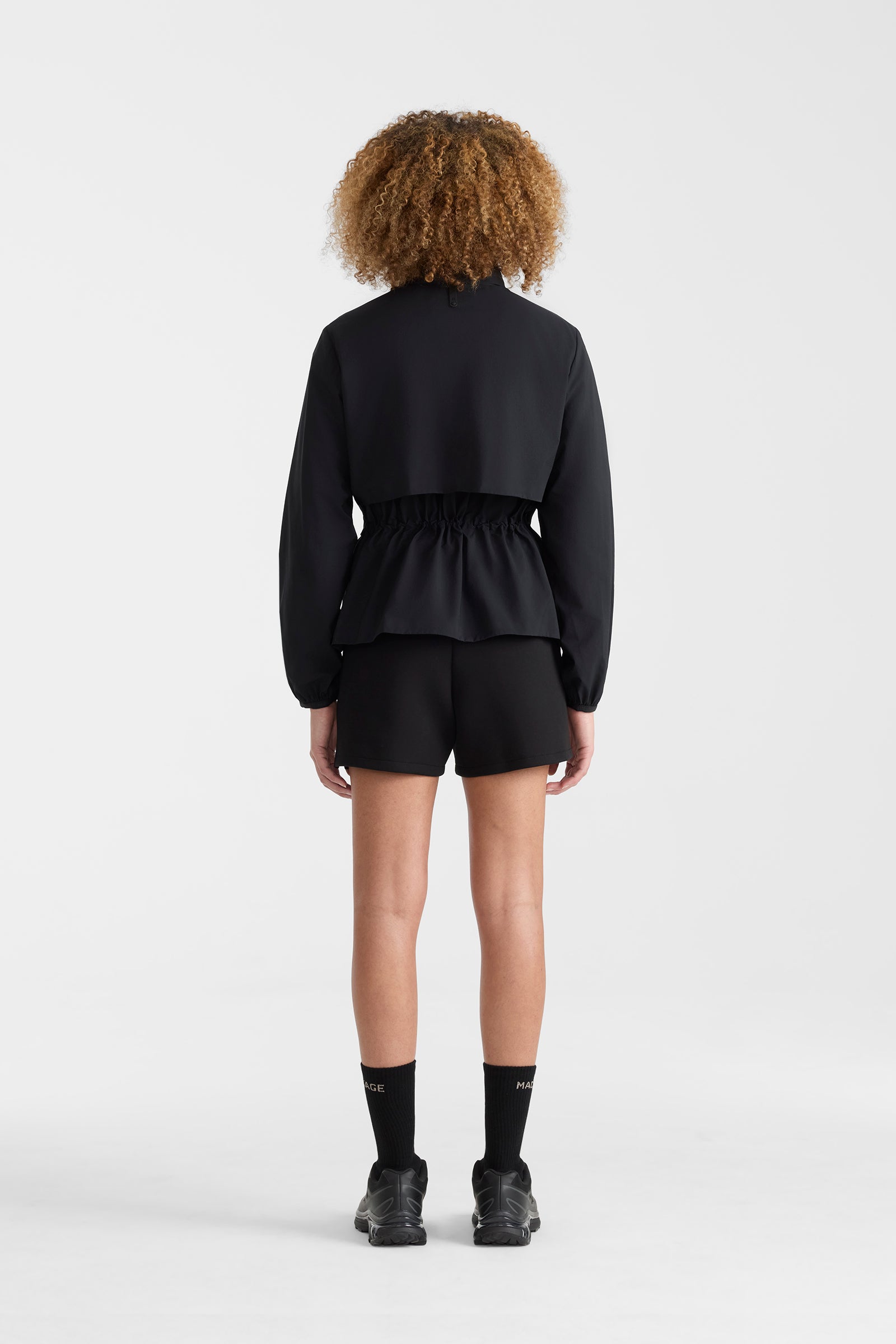 Woman wearing a black lightweight rain jacket with black shorts on a white background.
