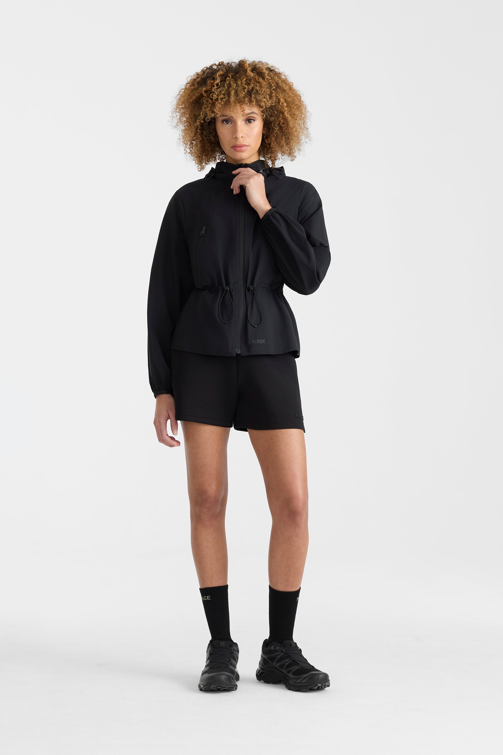 Woman wearing a black lightweight rain jacket with black shorts on a white background.