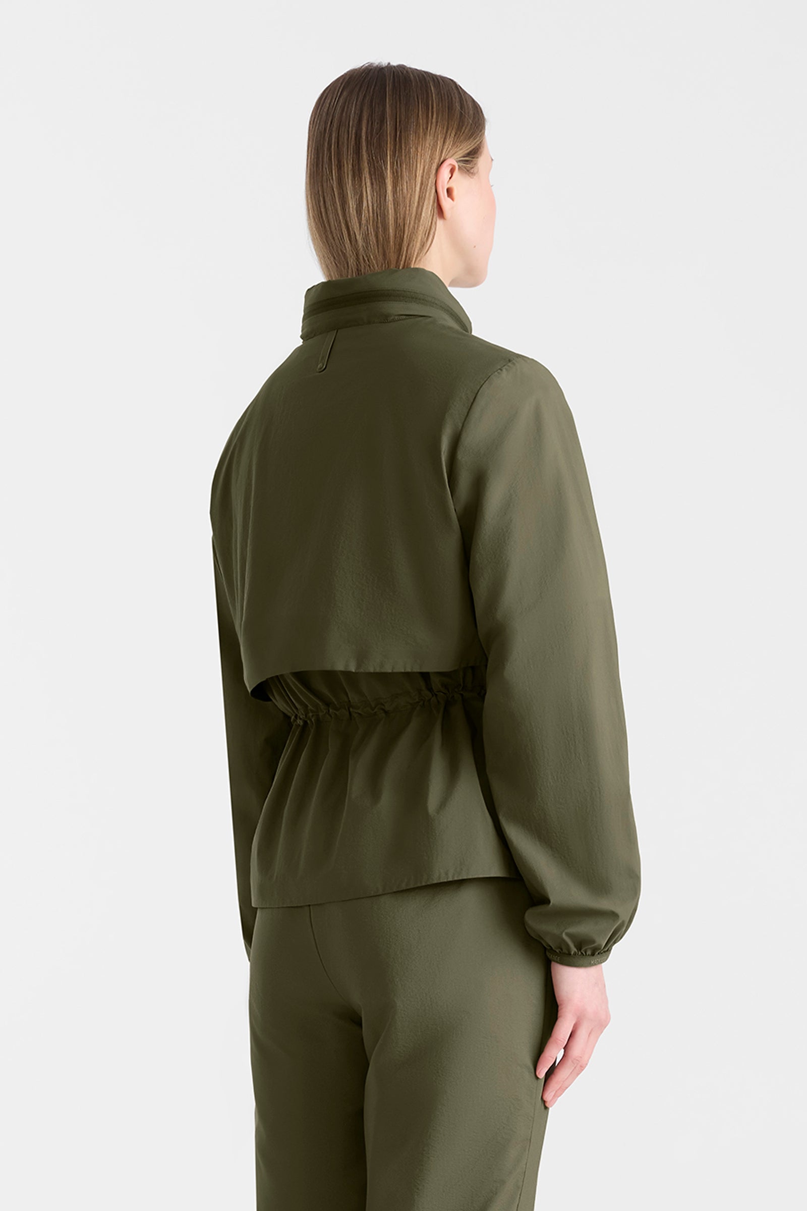 Woman wearing a light military jacket on and pants with a hood on a white background