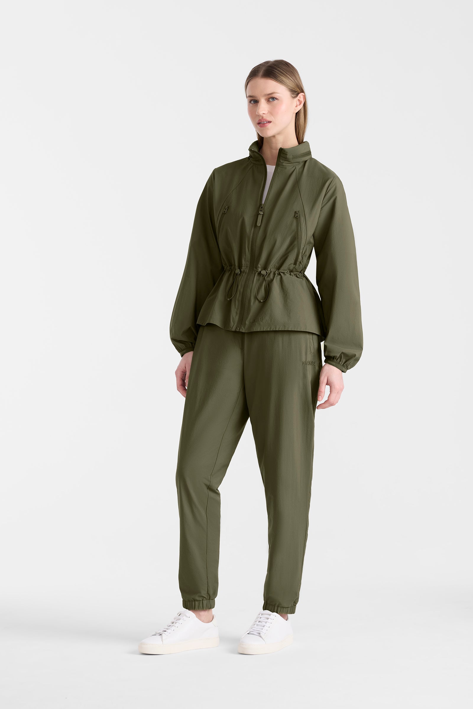 Woman wearing a light military jacket on and pants with a hood on a white background